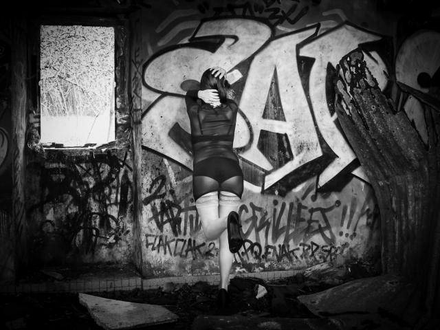 Fotografia in bianco e nero di BKLove con le mani sulla testa, che indossa una maglietta a maniche lunghe e pantaloncini, in piedi in una stanza fatiscente e ricoperta di graffiti, con una gamba sollevata, che mostra il suo sedere..