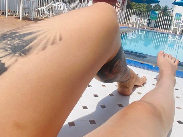 A coxa e a perna tatuadas de MelanyyLove estão em primeiro plano, com o pé descalço em uma piscina com piso de azulejos brancos. A tatuagem é um grande padrão de raios solares pretos..