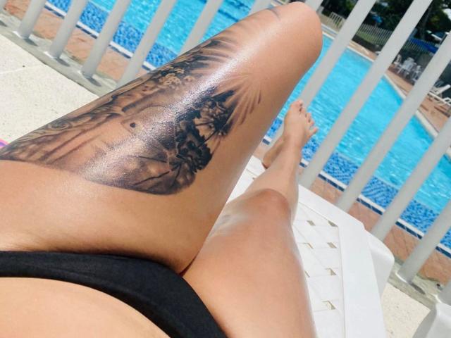 De glanzende, bruine benen van MelanyyLove, met een gedetailleerde zwarte tatoeage van een vrouw en bomen op haar rechter dij, liggen op een witte ligstoel bij een blauw zwembad..