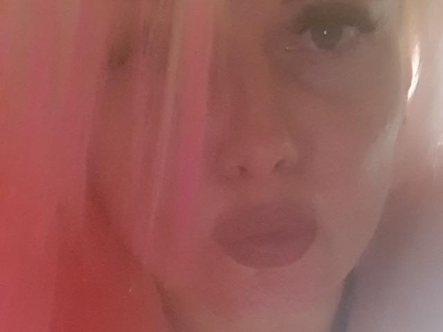 PinkDevil's face with pink hair covering one eye, pink lipstick, and slightly parted lips, blurred with soft pink and red light, creating a sensual, semi-transparent effect.