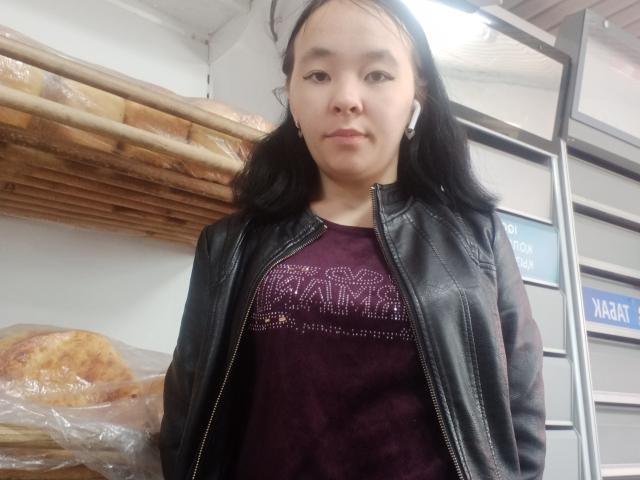 MalishkaSexy, uma mulher asiática de pele média, com cabelo preto, vestindo uma jaqueta de couro preta e uma camisa roxa, em pé em uma padaria com pães nas prateleiras..
