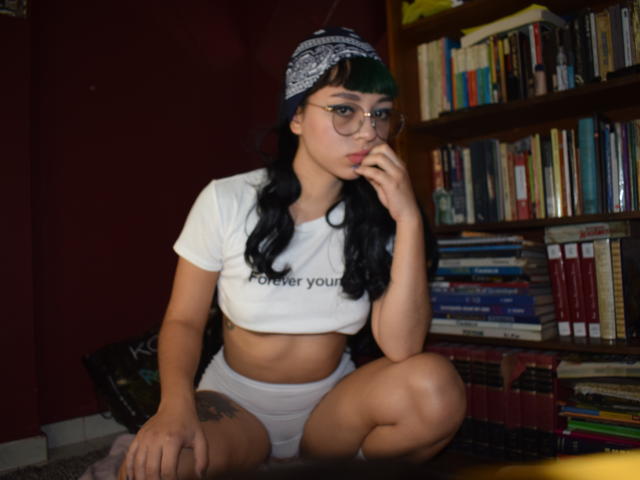 MefistoErica with green-tipped black hair, glasses, white crop top, white shorts, bandana, crouching, touching lips, tattoo on right thigh, bookshelf in background.