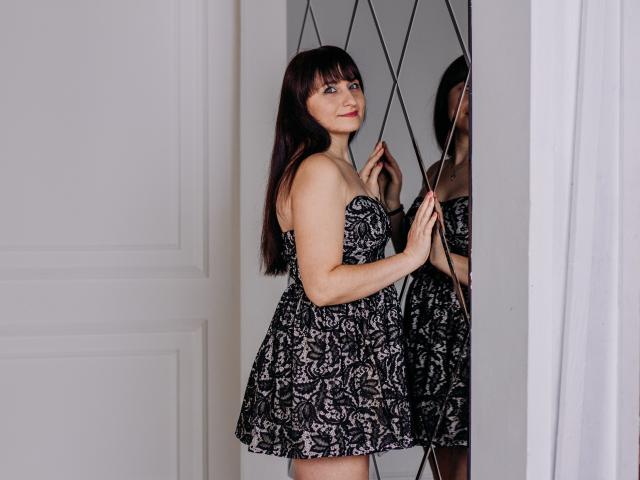 NinaFlawlees with dark brown hair, wearing a black and white floral strapless dress, standing next to a mirrored wall, smiling at her reflection, holding her dress's side.
