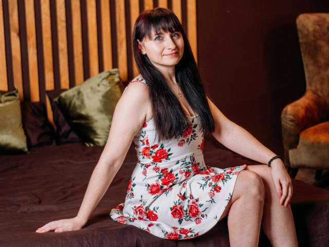 NinaFlawlees, with long black hair and bangs, sits on a bed in a white floral dress with red roses, showing her tits, wearing a black bracelet, smiling.