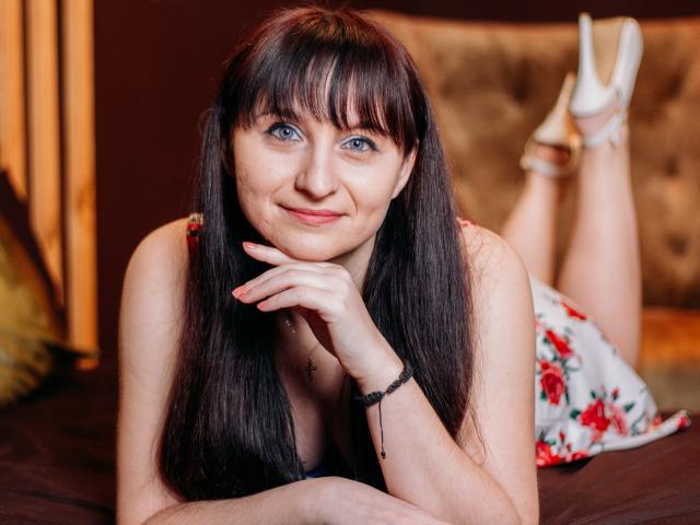 NinaFlawlees, lying on a bed, has long dark brown hair, blue eyes, and a cute smile. She's wearing a floral dress, white heels, and a black bracelet.