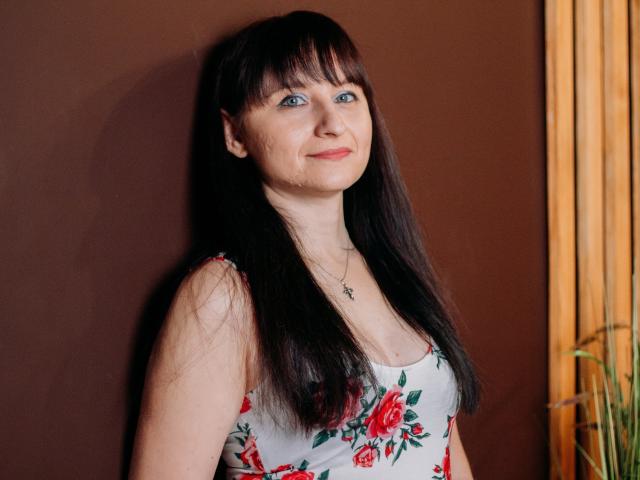 NinaFlawlees has long black hair, blue eyes, and a subtle smile. She's wearing a floral dress with red roses, showing her tits and wearing a necklace.