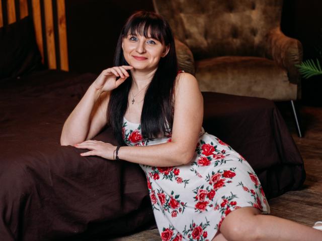 NinaFlawlees with long black hair, blue eyes, and a smile, wearing a floral dress, sitting on a brown bed, hand on chin, looking cute with a small ass.