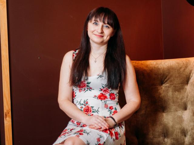 NinaFlawlees has long brown hair, blue eyes, and a small smile. She's wearing a floral dress, sitting on a beige chair, with her hands on her knees.