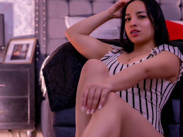 GiannaRusell, con senos de tamaño medio, sentada en una silla negra, vistiendo una blusa a rayas, con las piernas cruzadas, cabello negro, mano en la cabeza, sonrisa sutil, esmalte de uñas, collar, iluminación interior..