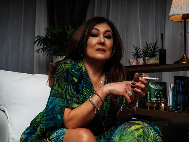 Freya Davidson, con cabello castaño oscuro, un vestido de color verde con estampado de hojas, esmalte de uñas negro, sosteniendo un cigarrillo, pulseras de plata, sentada en una silla blanca, con una expresión segura, de busto medio, con una piel ligeramente bronceada..