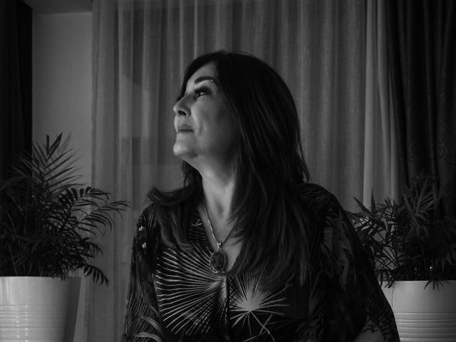 Freya Davidson, com cabelo escuro, olhando para cima, vestindo uma camisa estampada, com um colar com um pingente redondo, sentada em frente a plantas em vasos em uma foto em preto e branco..