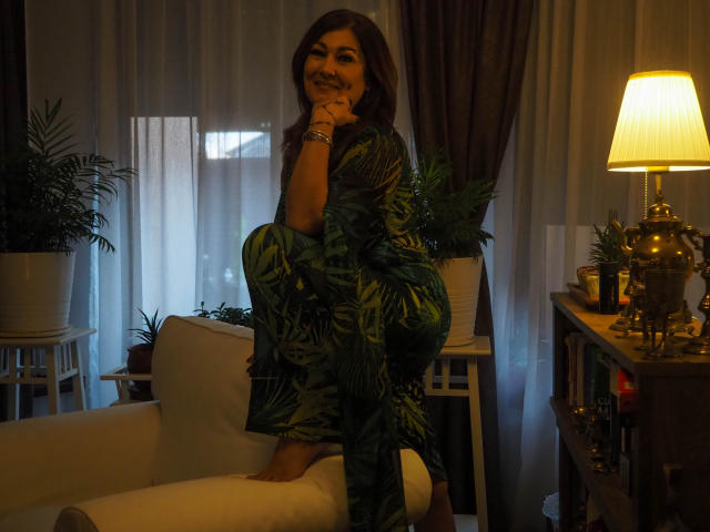 Freya Davidson, con un vestido de color marrón con estampado de hojas verdes y cabello castaño, está sentada en una silla blanca, sonriendo, con la mano en la barbilla, en una habitación con poca luz, con una lámpara a la derecha y plantas a la izquierda y a la derecha..