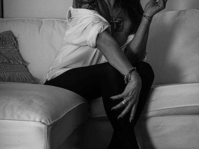 Freya Davidson, com os seios parcialmente expostos, vestindo uma camisa larga, calças pretas e joias, sentada em um sofá branco, levantando a mão esquerda. Foto em preto e branco..
