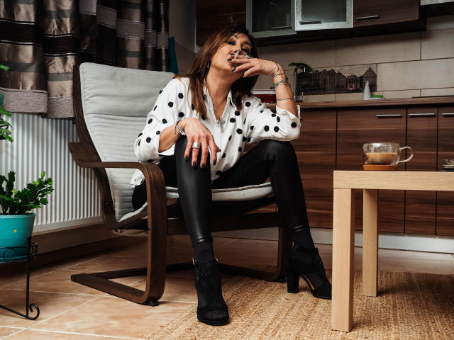 Freya Davidson, con cabello castaño, una blusa blanca con lunares, pantalones de cuero negro y botas de tacón alto, bebiendo de una taza, sentada en una silla de madera en una cocina..