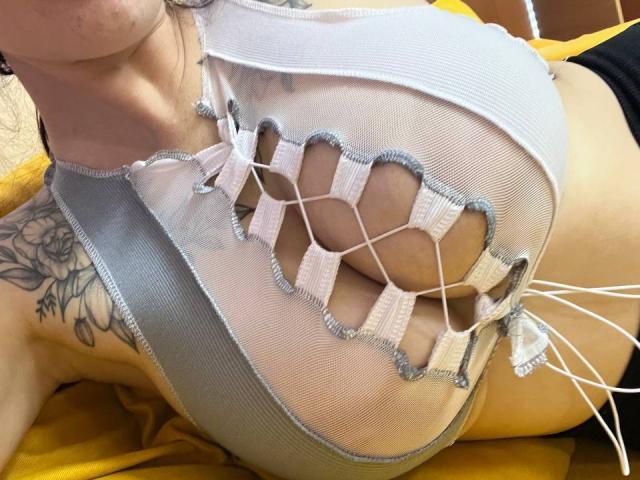 Les grandes seins d'Innysik sont visibles dans un haut blanc en dentelle et en mesh, qui révèle sa poitrine et ses tatouages sur le côté droit, alors qu'elle est allongée sur une surface jaune. Son visage est partiellement visible..