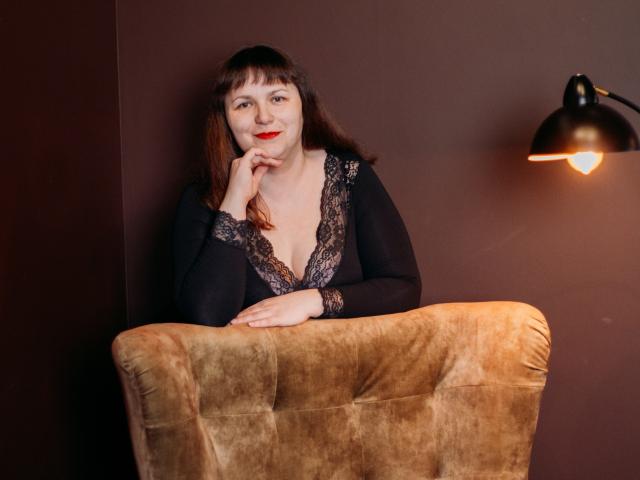 AliceInks with red lipstick, black lace top, and brown hair, leaning on a tan suede chair, smiling with one hand on her chin, under a black wall lamp.