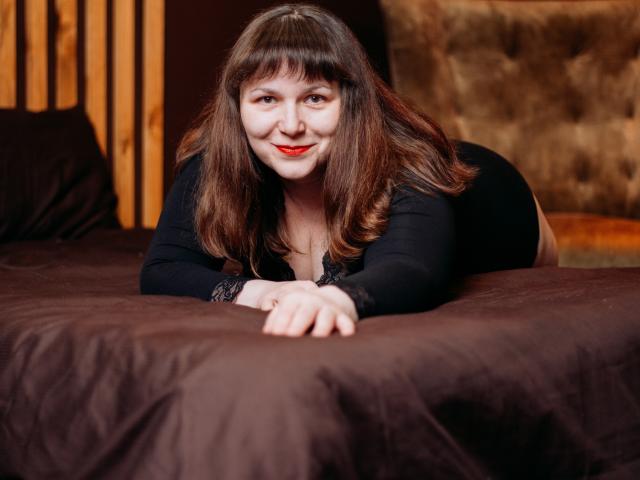 AliceInks, with red lipstick and black lace sleeves, lies on a brown bed, smiling at the camera, her ass up, hands in front, brown hair, wooden headboard.