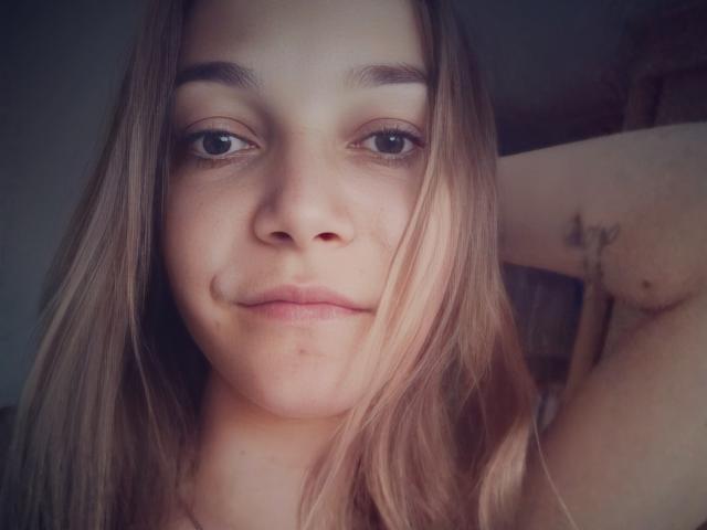 LovelyxEmilly has light brown, straight hair, a small, cute nose, and a slight smile. She's looking at the camera with brown eyes, and a tattoo is visible on her arm.