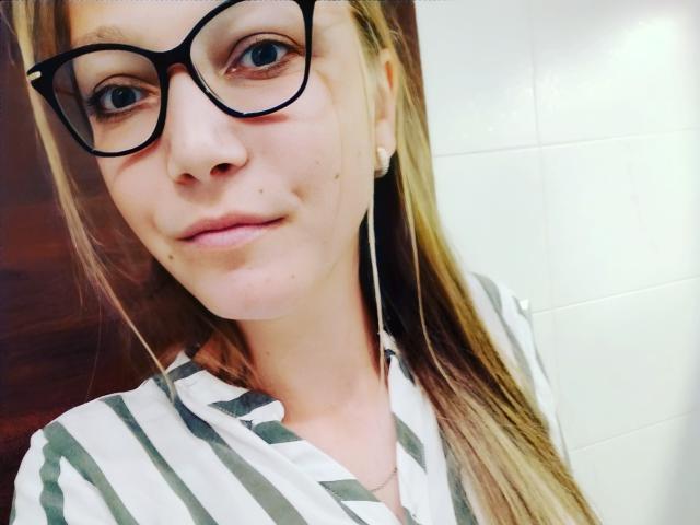 LovelyxEmilly con gafas, cabello rubio y una camisa a rayas, mostrando una pequeña peca en su mejilla derecha y una sonrisa sutil. Pared de azulejos blancos en el fondo..