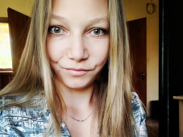 LovelyxEmilly has blonde hair, green eyes, and a small mole under her right eye. She's wearing a patterned shirt and a silver necklace, smiling slightly.