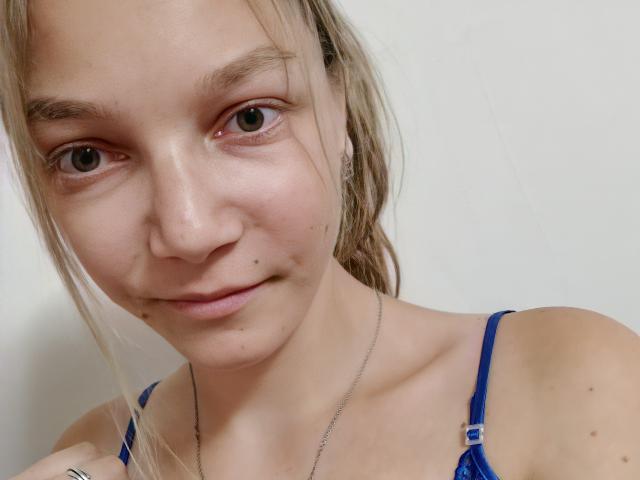 LovelyxEmilly with light blonde hair, blue eyes, and a small mole on her right cheek, wearing a blue bra, and a silver necklace, in a soft, natural light.