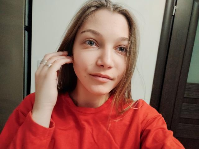 LovelyxEmilly with light brown hair, blue eyes, wearing a red shirt, hand touching ear with ring, soft smile, subtle makeup, slightly freckled skin, casual selfie.