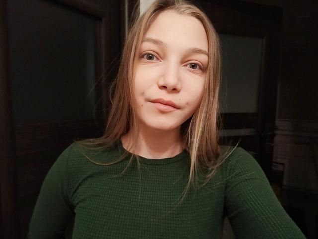 LovelyxEmilly with blonde hair, green eyes, and a small beauty mark on her upper lip, wearing a green rib-knit top, smiling softly in a dimly lit room.