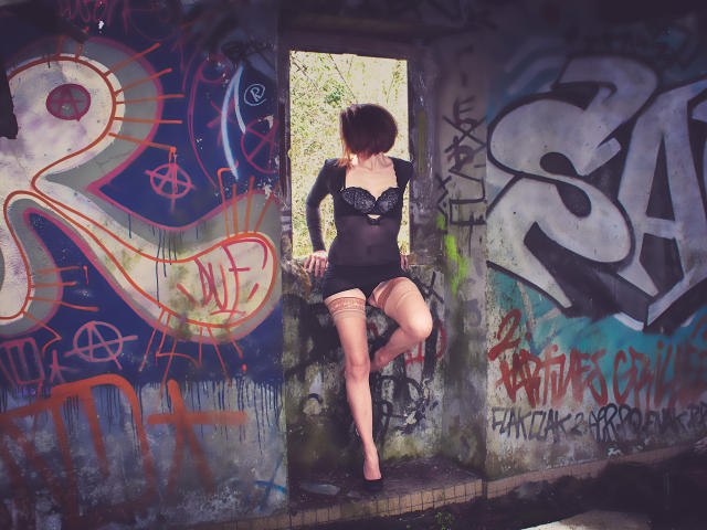 BKLove, con un sedere prosperoso, indossa lingerie nera e calze, ed è appoggiata a una finestra coperta di graffiti, con una gamba sollevata, in un ambiente urbano degradato. Il suo seno è piccolo, coperto..