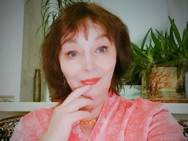 MilaMils mit kurzen, braunen Haaren, blauen Augen und rotem Lippenstift, trägt ein pinkes, geblümtes Hemd, berührt ihr Kinn, lächelt und trägt eine Perlenkette..