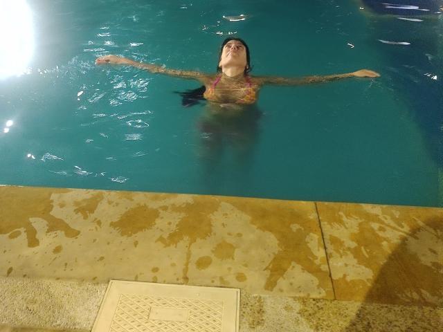 SeleneBrigth in a pool with arms outstretched, wearing a colorful bikini, her tattooed arms and dark hair visible, her eyes closed, floating in clear blue water.