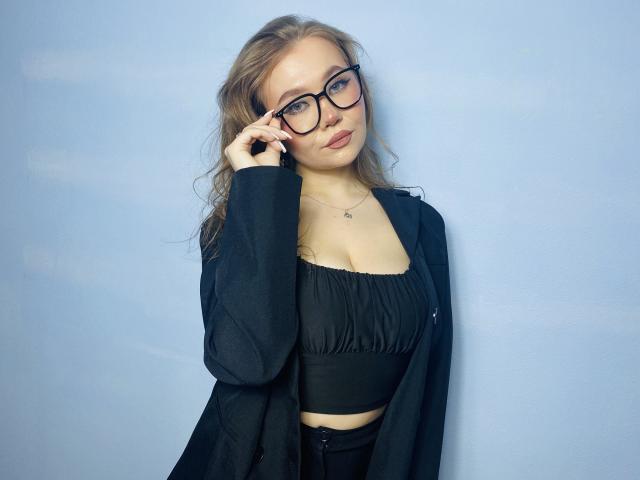 Stacy Yellig, con gli occhiali, capelli biondi ondulati, top nero che mostra il seno, giacca nera e pantaloni neri, tiene il telefono all'orecchio, sfondo blu..