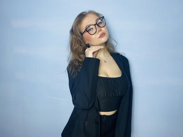 StacyYellig with wavy brown hair, glasses, black crop top showing her tits, and black pants, wearing a black blazer, hand on neck, looking down, against a light blue wall.