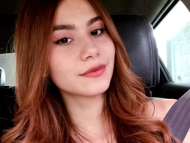 CiaraForest tiene sombra de ojos rosa claro, cabello ondulado castaño y lápiz labial rosa, y está sentada en un coche con un asiento de cuero negro detrás de ella. Sus senos no son visibles..