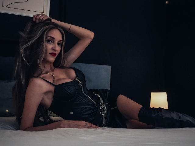 MaiaMoxie, avec de longs cheveux blonds, du rouge à lèvres rouge, une robe en cuir noir et des bottes, allongée sur un lit, une main dans les cheveux, sexy, mignonne, petits seins, lit blanc, éclairage tamisé..
