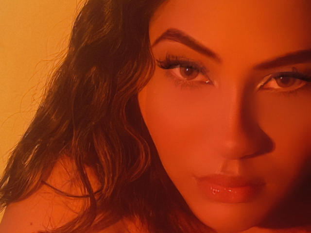 KataMillerr's face in a warm orange light, with wavy brown hair, thick eyebrows, long eyelashes, and glossy pink lips, staring directly into the camera. Boobs not visible.