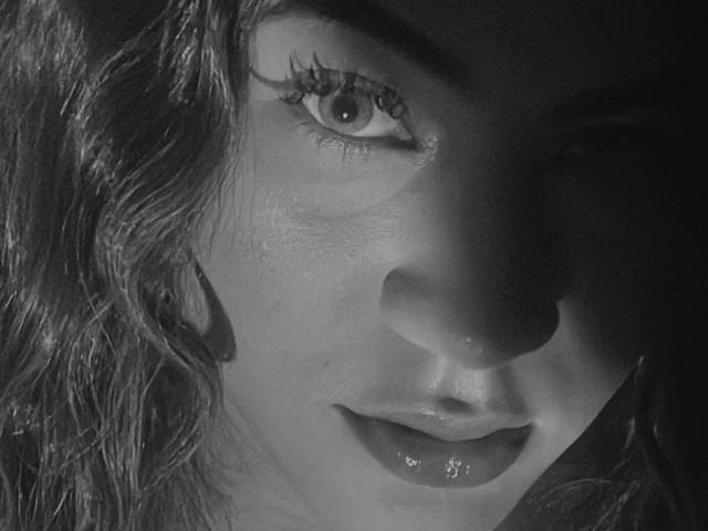 Le visage de KataMillerr en noir et blanc, montrant ses lèvres pulpeuses, ses cheveux ondulés et ses longs cils. Son œil droit est net, tandis que le côté gauche est ombragé..