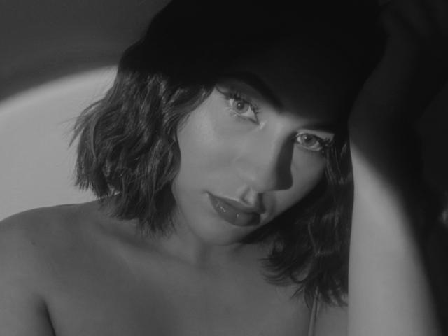 Photo en noir et blanc de KataMillerr, avec des cheveux courts et ondulés, regardant vers le haut avec des yeux séduisants, des lèvres foncées et une ombre sur son visage, laissant apparaître ses épaules nues et un aperçu de ses seins..