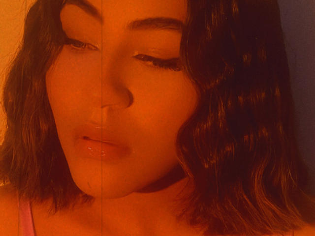Le visage de KataMillerr est partiellement divisé par une ligne verticale, avec un éclairage orange chaud. On peut voir ses yeux à moitié fermés, ses cheveux bruns ondulés et ses lèvres brillantes. Son haut rose est discret..