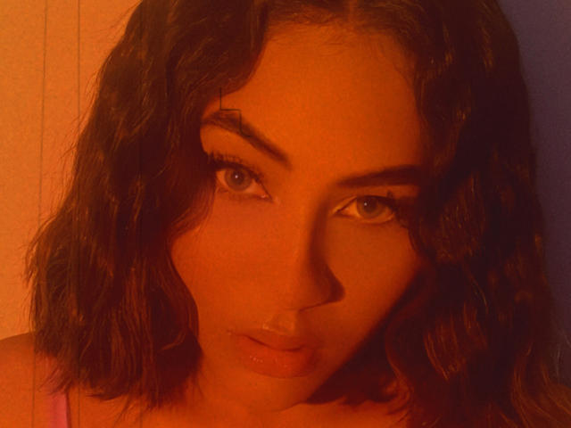 KataMillerr with wavy brown hair, orange-tinted skin, and serious eyes, looking directly at the camera, showing a slight pout and small nose ring. Warm, dim lighting.