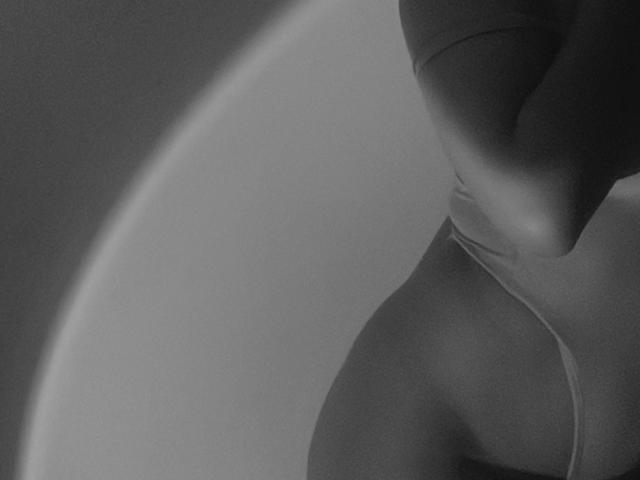 KataMillerr's upper body, grayscale, with soft lighting, showing a curve of her tits and a strap down her side, creating a sensual, shadowy, partial profile.