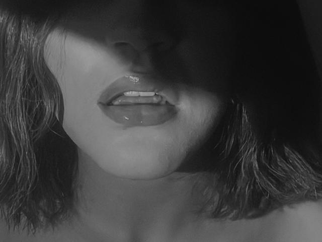 KataMillerr's glossy lips are slightly parted, showing a hint of teeth, with dark shadows over her face and shoulder-length, wet-looking hair in a black-and-white photo.