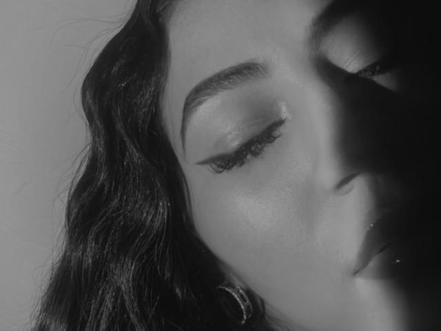 KataMillerr's face in black-and-white, eyes closed, with subtle eyeliner, dark wavy hair, and a small hoop earring on the left side. Soft shadows on her cheek.