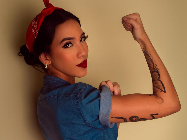 KylieWhite with red bandana, blue denim shirt, raised arm, tattoo on forearm, dark lipstick, earrings, looking back, smiling, showcasing her arm flex. Her tits aren't visible.