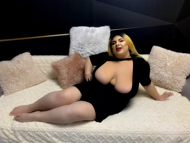 HornyBbw met grote borsten, liggend op een bed, gekleed in een zwarte jurk, blond haar, rode lippenstift, een witte, geribbelde deken, roze, pluizige kussens, een zwarte en gouden muur op de achtergrond..