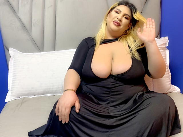 HornyBbw met grote borsten in een zwarte jurk, blond haar, zwaaiend, zittend op een bed, blauwe muren, grijze hoofdbord, witte kussens, roze kussen, gouden armband, kijkend naar de camera..