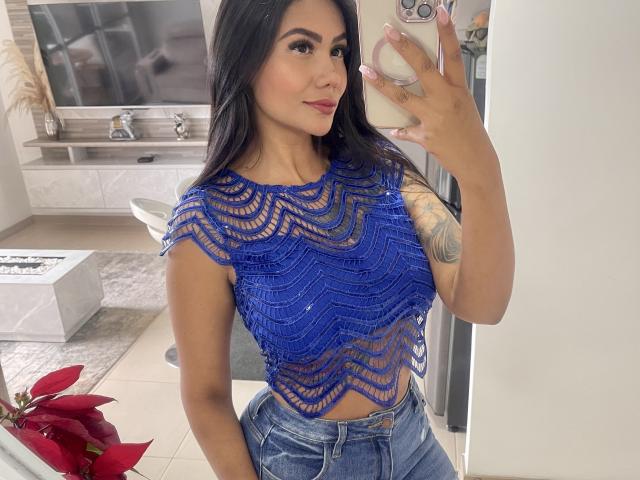 AlessandraMalory with dark hair, wearing a blue, wavy-patterned, see-through top, showing her tits, and high-waisted jeans, taking a mirror selfie, showing a tattoo on her arm.