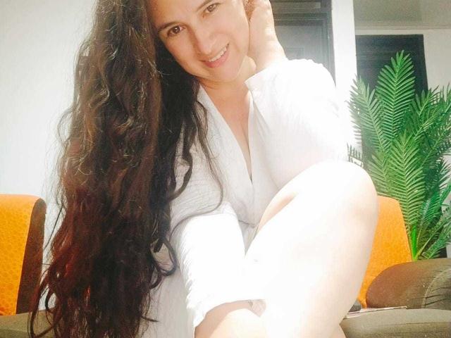 Brihana Charlot, com cabelos longos, ondulados e castanho-escuros, sorrindo, vestindo uma camiseta branca texturizada, sentada com as pernas cruzadas, exibindo seu traseiro claro, curvilíneo e nu em um quarto bem iluminado..