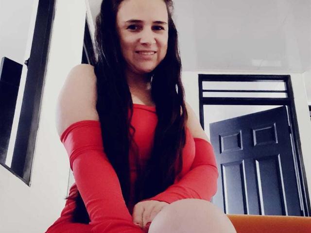 Brihana Charlot, con el pelo largo y oscuro, está sentada sonriendo, vestida con un vestido rojo de un solo hombro, que deja al descubierto sus hombros y brazos, y donde se pueden ver ligeramente sus senos, en una habitación muy iluminada..