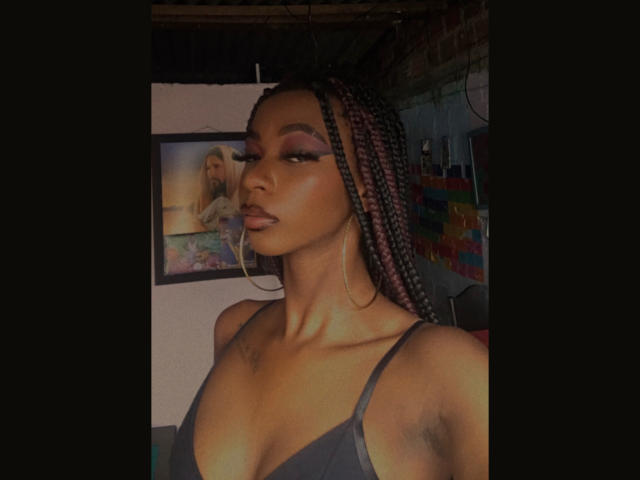 Keirysex with dark skin, braided pink hair, big hoop earrings, and black top showing her tits, has bold makeup and a confident expression. Her armpits are slightly visible.