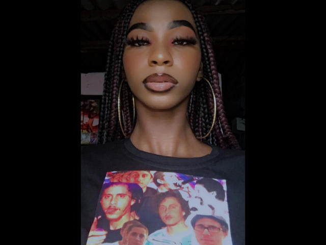 Keirysex tiene la piel oscura, labios carnosos y cabello trenzado. Lleva grandes aros y una camisa negra con una foto colorida de personas en la parte delantera..
